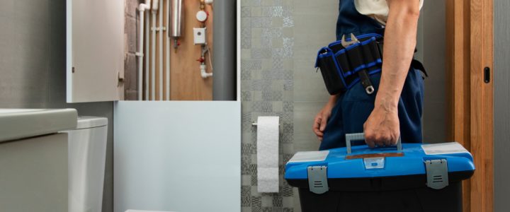 Problemas de sucção do vaso sanitário: o que causa? Como resolver? Problema de ventilação: encanador de jardineira jeans e blusa branca com uma maleta de ferramentas na mão esquerda de frente para um vaso branco em um banheiro com azulejo cinza