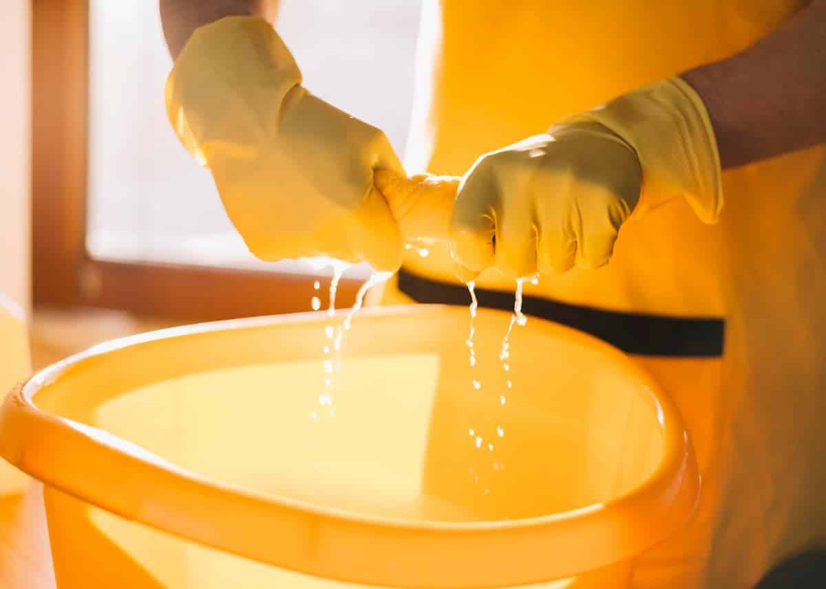 mãos com luvas amarelas torcendo um pano cheio de água sobre balde