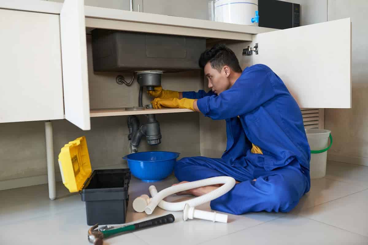 profissional desentupidor sentado no chão da cozinha mexendo em cano embaixo da pia