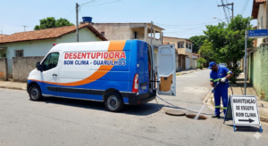 Unidade móvel da Desentupidora Bom Clima estacionada em uma rua residencial de Guarulhos. Um técnico uniformizado realiza o hidrojateamento em um bueiro aberto, utilizando equipamentos de alta pressão. Ao lado, uma placa de sinalização indica 'Manutenção de Esgoto Bom Clima'